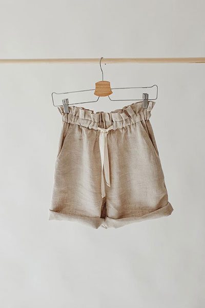 Tan linen shorts with a gathered waist, on a hanger against a white wall.