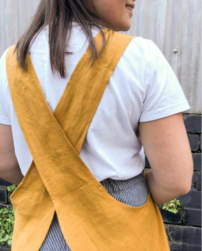 A mustard linen cross-back apron worn over a white t-shirt.