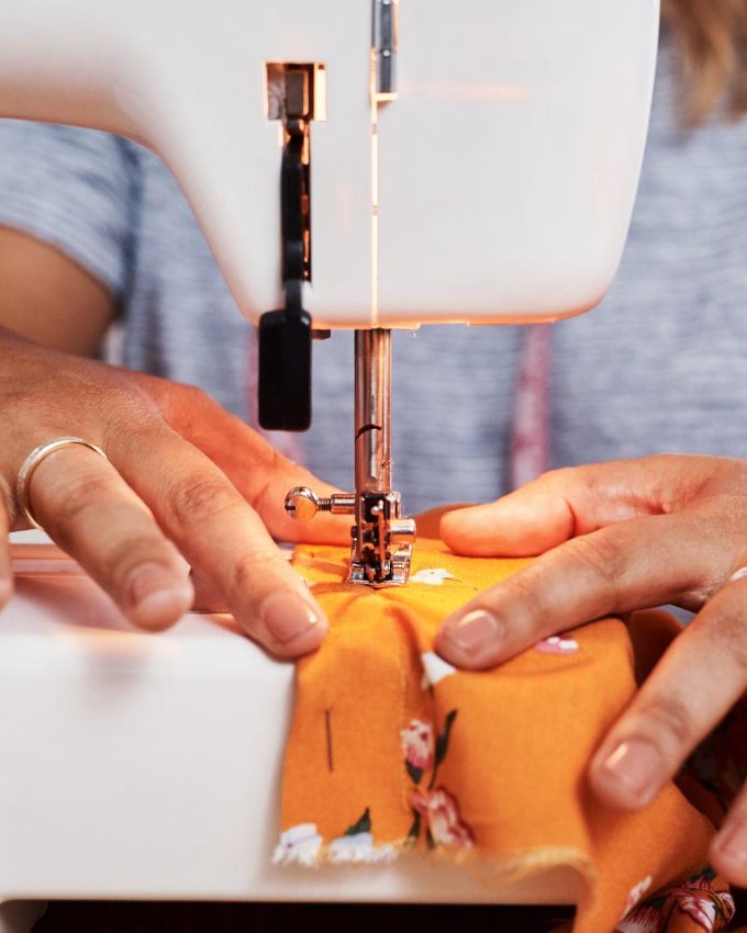 Hands guiding orange floral fabric through a sewing machine.