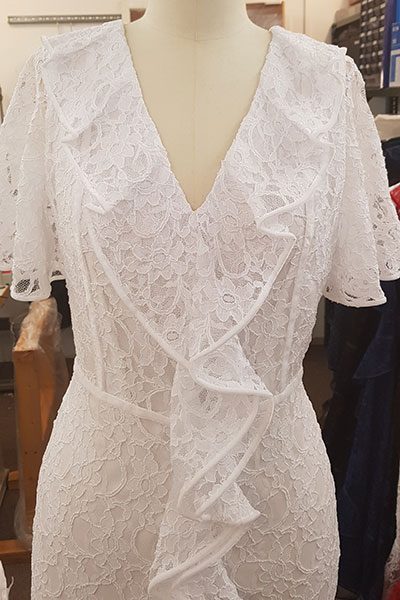 A white corded-lace dress with a ruffled neckline on a dress stand.