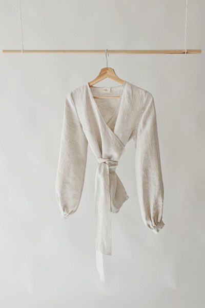 A cream linen wrap top with a tie fastening, hanging from a wooden rail.